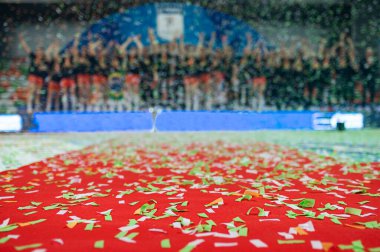 Kırmızı halıda konfeti ve arka planda spor takımları.