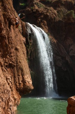 Faslı genç bir adam yüksek kayalıklardan bir şelalenin altındaki suya atlıyor..
