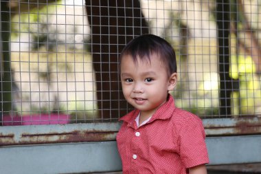 Kırmızı tişörtlü Asyalı bir çocuk kuş kafesinin yanında mutlu bir şekilde gülümsüyor..