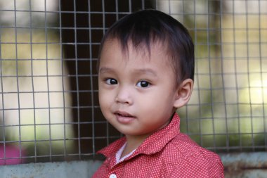 Kırmızı tişörtlü Asyalı bir çocuk kuş kafesinin yanında mutlu bir şekilde gülümsüyor..