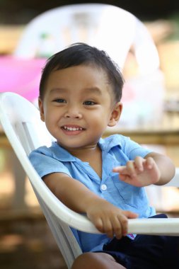 Mavi gömlekli Asyalı bir çocuk beyaz bir sandalyede oturuyor, mutlu bir şekilde gülümsüyor..