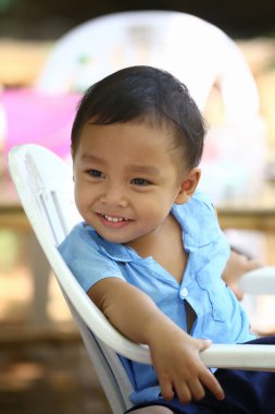 Mavi gömlekli Asyalı bir çocuk beyaz bir sandalyede oturuyor, mutlu bir şekilde gülümsüyor..