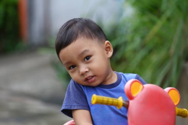 İki yaşlarında Asyalı bir çocuk mavi bir gömlek giyiyordu. Oyuncak arabasının üzerinde oturuyordu ve mutlu bir şekilde gülümsüyordu..