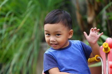 İki yaşlarında Asyalı bir çocuk mavi bir gömlek giyiyordu. Oyuncak arabasının üzerinde oturuyordu ve mutlu bir şekilde gülümsüyordu..