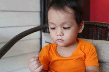 İki yaşlarında, turuncu gömlekli Asyalı bir çocuk sandalyeye oturmuş elinde bir şeye tuhaf bir şekilde bakıyordu..