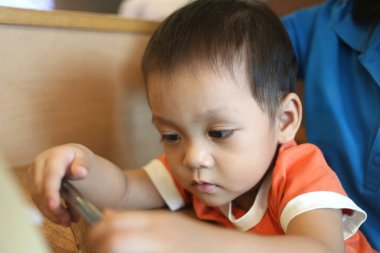 Asyalı çocuk akıllı telefona bakıyor, arkasında annesi olan bir şeye dikkat çekiyor..