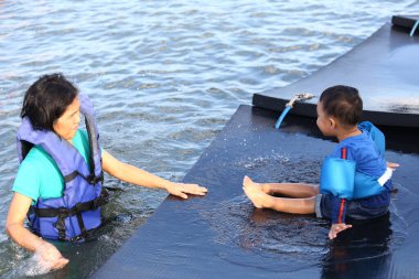 Asian mother playing with her son on a raft in the sea, both wearing life jackets to prevent drowning. clipart