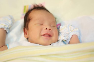 Yeni doğmuş bir bebek beyaz bir yatakta uyuyor ve mutlu bir şekilde gülümsüyor, eldiven giyiyor ve beyaz-sarı bir battaniyeyle örtülü..