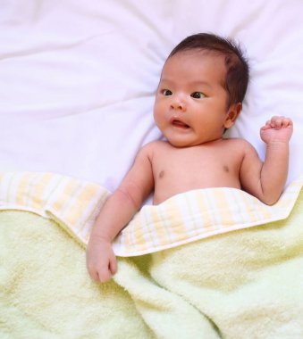 Küçük bir çocuk, beyaz bir yatak ve sarı bir battaniye üzerinde şaşkın şaşkın yatıyor..