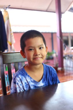 Neşeli genç bir çocuk sıcak bir gülümseme sergileyerek bir masada oturur. Hareketli gömleği ve rahat duruşu ev dekoruna eğlenceli bir dokunuş katıyor..