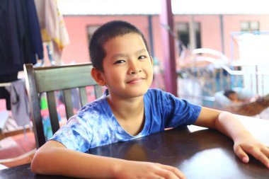 Genç bir çocuk parlak ve davetkar bir iç mekanda bir masada otururken gülümsüyor. Neşeli ifadesi çocukluk sevincini ve masumiyetini yansıtıyor..