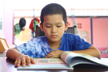 Genç bir çocuk bir masada oturuyor, renkli bir kitap okumaya derinden bağlıdır. İçerideki aydınlık ortam merak ve öğrenmeyi teşvik ediyor..