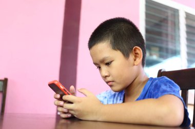 Genç bir çocuk, canlı bir kapalı mekan ortamında akıllı telefonuyla derinden meşgul bir şekilde masada oturuyor. Görüntü, gençlik ve teknoloji arasındaki bağlantıyı yakalıyor..