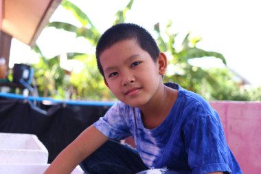 Mavi kravat boyalı gömlekli genç bir çocuk yeşilliğin ortasında otururken gülümsüyor, çocukluk sevincinin ve kaygısız maceraların ruhunu yakalıyor..