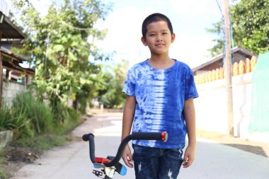 Güneşli bir günde bisikletinin yanında duran genç bir çocuk güven ve neşe saçıyor. Canlı mavi tişört, açık hava manzarasına renk katıyor..