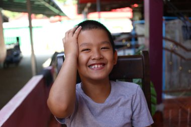 Neşeli bir çocuk neşeli bir gülümseme ile dışarıda oturuyor, oyun oynayarak elini başının üzerinde dinlendiriyor. Samimi an, çocukluk mutluluğunun özünü yakalar..