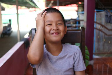 Mutlu genç bir çocuk dışarıda otururken parlak bir şekilde gülümser. Neşeli ifadesi çocukluk ve kaygısız anların özünü yakalıyor, canlı renklerle çevrili..