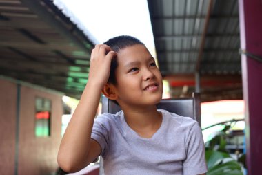 Genç bir çocuk düşünceli bir şekilde oturur, bir tebessümle çeşitli duygular gösterir. Günlük kıyafetleri, bir iç mekan ortamında rahatlamış bir düşünce anını yansıtıyor..