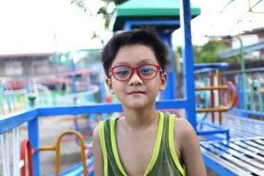 Gözlük takan genç bir çocuk canlı bir oyun sahasında kendinden emin duruyor. Renkli ekipmanlar, çocukluk oyununun özünü vurgulayarak neşeli atmosferi güçlendirir..