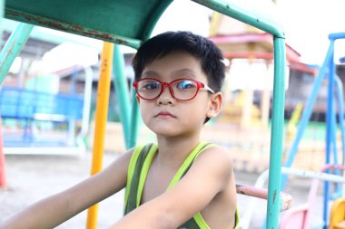 Kırmızı gözlüklü genç bir çocuk canlı bir oyun alanında eğleniyor. Bu samimi an çocukluk oyununun neşesini ve enerjisini yakalar..