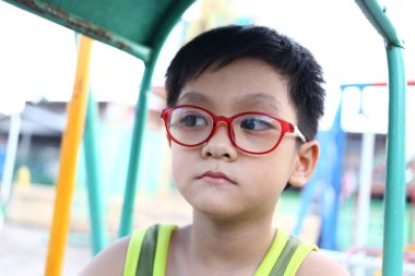 Parlak kırmızı gözlük takan düşünceli genç bir çocuk oyun parkında oturuyor. Görüntü, çocukluk masumiyetini ve merakını canlı bir ortamda gösteren bir iç gözlem anı yakalıyor..