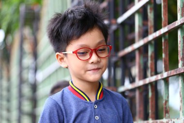 Kırmızı gözlüklü neşeli genç bir çocuk açık havada güneşli bir günün tadını çıkarırken gülümsüyor, neşeli kişiliğini ve şık görünümünü sergiliyor..