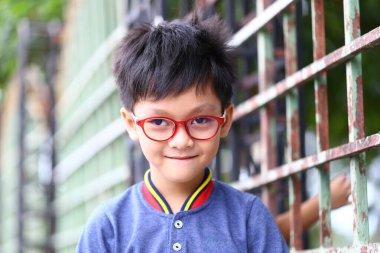 Gözlüklü neşeli bir çocuk yeşil bir çitin yanında güvenle gülümsüyor. Neşeli ifadesi gençliğin ve masumiyetin özünü canlı bir ortamda yakalıyor..