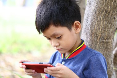 Genç bir çocuk bir ağacın altında oturuyor, akıllı telefonuna odaklanmış, sakin bir ortamda doğa ve teknolojinin kesişimini gösteriyor..