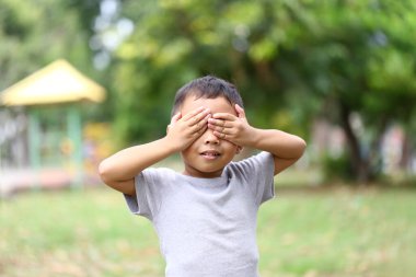 Neşeli bir çocuk ellerini kullanarak yeşil bir parkta saklambaç oynuyor. Bu an çocukluk masumiyetinin ve keşiflerin özünü yakalar..
