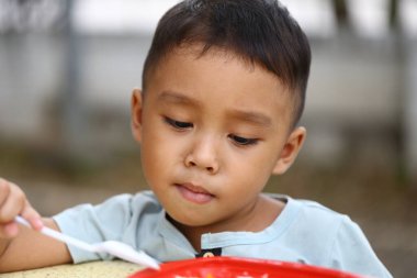 Küçük bir çocuk yemek yerken konsantrasyon ve neşe gösteren kaşıkla yemekten zevk alır. Bu portre çocukluğun masumiyetini ve mutluluğunu yansıtır..