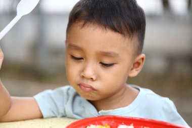 Genç bir çocuk düşünceli bir şekilde dışarıda oturuyor, tabakta renkli bir yemeğin tadını çıkarıyor. Bu ifade, çocukluğun huzurlu bir anını yakalayan merak ve neşeyi yansıtır..