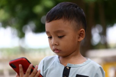 Genç bir çocuk açık havada akıllı bir telefonla iletişim kuruyor, merakını ve dikkatini gösteriyor. Bu görüntü çocukluk ve teknolojinin doğal ortamını yansıtır..