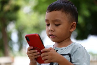 Genç bir çocuk yoğun bir şekilde açık hava ortamında akıllı telefonuna odaklanıyor, modern teknolojiyle olan ilgisini ve ilgisini somutlaştırıyor..