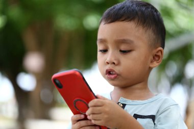 Açık havada akıllı telefonuyla meşgul olan genç bir çocuk merakını ve dikkatini gösteriyor. Canlı doğal ortam çocukluğun neşeli atmosferini güçlendirir..