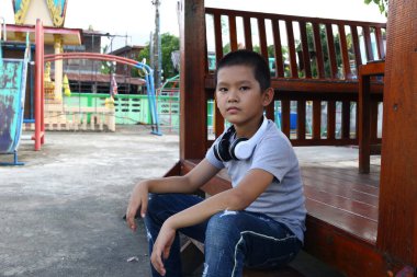 Kulaklıklı genç bir çocuk oyun parkındaki ahşap bir bankta rahat rahat oturur, açık havada, canlı bir ortamda eğlenir. Yaz ve çocukluk temaları için mükemmel.