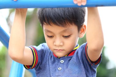 Genç bir çocuk oyun parkı ekipmanlarına tutunuyor, açık hava aktiviteleri sırasında konsantrasyon ve neşe sergiliyor. Bu resim çocukluk oyunlarını ve keşifleri gün ışığında yakalar..