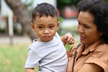Bir anne, genç oğlunu parkta tutarak ailenin mutluluğunu ve açık hava anlarının güzelliğini kutlar. İfadeleri aşkı ve birlikteliği yansıtıyor..