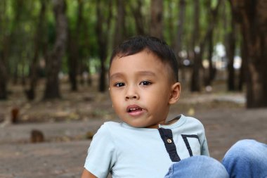 Genç bir çocuk, ağaçlarla çevrili sakin bir orman ortamında yerde oturuyor. Onun sakin ifadesi doğadaki masumiyet ve neşeyi yakalar..