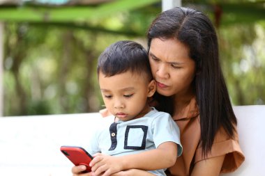 Bir anne ve küçük çocuğu açık havada bir anı paylaşıyor, akıllı telefona bakıyor. Sahne, modern ebeveynliğin ve teknolojinin özünü güzel bir doğal ortamda yakalıyor..