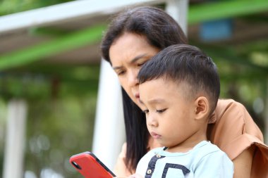 Bir anne ve küçük çocuğu açık havada bir anı paylaşıyor, akıllı telefona odaklanıyor. Bu sahne modern ebeveynliğin ve teknoloji kullanımının özünü yansıtır..