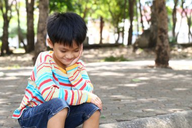 Genç bir çocuk düşünceli bir şekilde bir parkta oturur ve etrafındaki doğanın tadını çıkarır. Renkli gömleği, güneşli bir günün neşeli atmosferine renk katıyor..