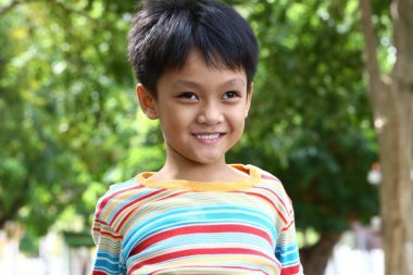 Neşeli genç bir çocuk renkli çizgili bir gömlek giyerken parlak bir şekilde gülümsüyor. Canlı yeşilliklerle çevrili, mutluluğu ve tasasız çocukluk ruhunu temsil ediyor..