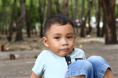 Genç bir çocuk sakin bir doğa parkında oturuyor ve tuhaf bir ifade sergiliyor. Ağaçlar ve güneş ışığıyla çevrili bu resim çocukluk masumiyetini ve neşesini yakalar..