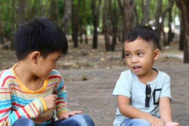 İki genç çocuk, doğa ve ağaçlarla çevrili bir parkta eğlenceli bir anı paylaşıyor. İfadeleri kaygısız bir ortamda dostluk ve neşe taşır..