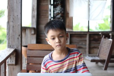 Kırsal bir yerde oturan genç bir çocuk, kulaklık takarken dizüstü bilgisayara odaklanmış. Sıcak ahşap çevreler öğrenmek için sakin bir atmosfer yaratır..