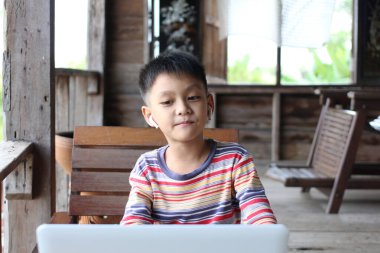 Genç bir çocuk rahat bir ahşap ortamda otururken dizüstü bilgisayar kullanmaktan hoşlanır. Rahatlamış tavrı ve kulaklığı bu neşeli ortamda öğrenme deneyimini arttırıyor..