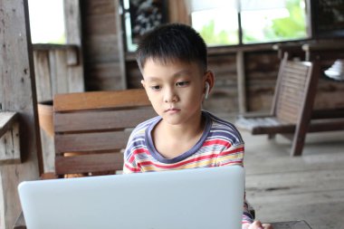 A young boy sits in a rustic interior, focused on his laptop while wearing headphones. The cozy setting highlights his engagement with technology and learning. clipart