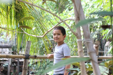 Neşeli genç bir çocuk bambu bir korkuluğun yanında duruyor canlı yeşil bir ortamda, yemyeşil yapraklarla çevrili, doğada neşe ve masumiyet saçıyor..