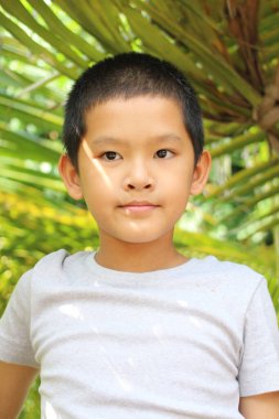 Genç bir çocuk açık havada duruyor, canlı yeşillikler arasında nazik bir gülümseme sergiliyor. Yumuşak güneş ışığı onun neşeli ifadesine sıcaklık katar, masumiyeti yakalar..