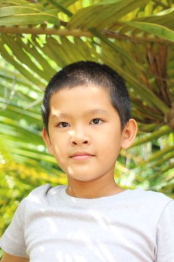 Genç bir çocuk yemyeşil bir zemine karşı duruyor, kendine güven ve masumiyet saçıyor. Güneş ışığı yaprakların arasından süzülerek sakin bir atmosfer yaratır..